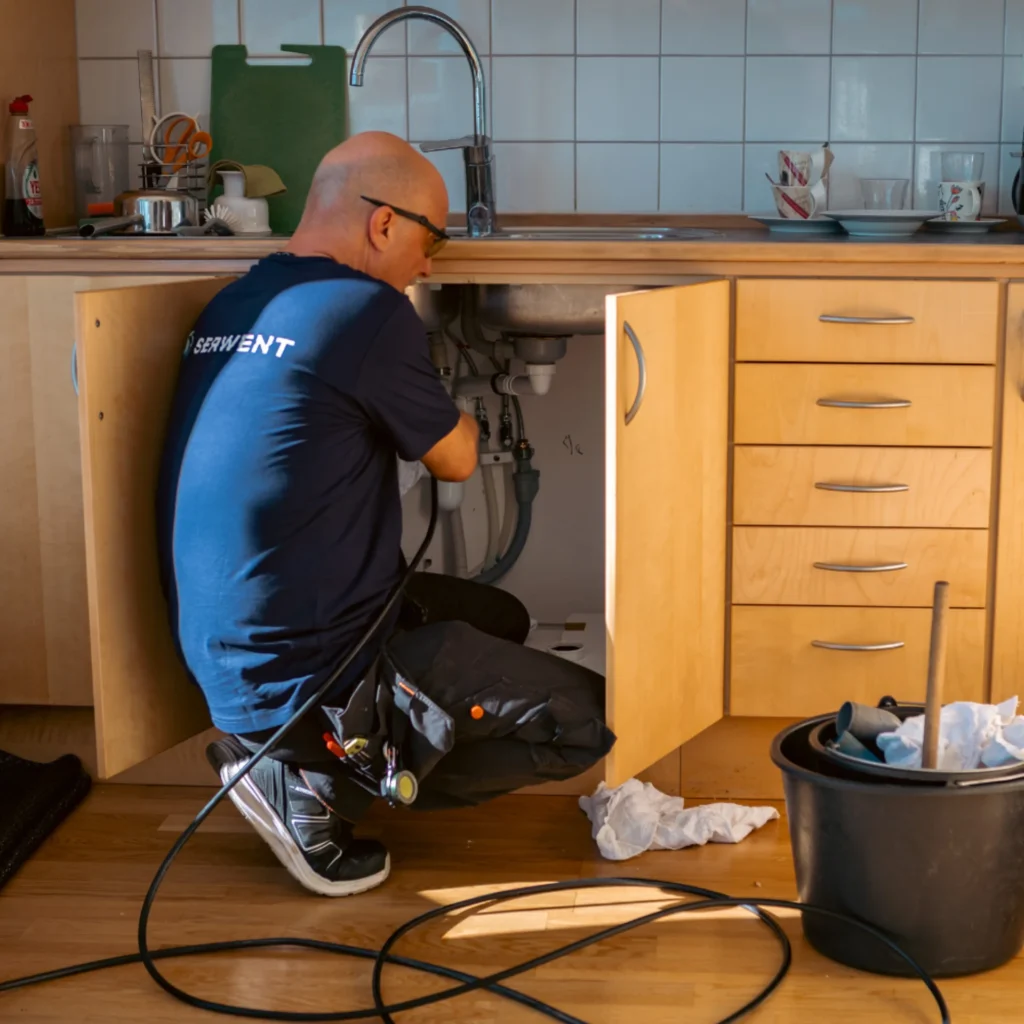 En rörmokare i marinblå skjorta och glasögon står på knä under en köksbänk med öppna skåpluckor och arbetar med rören - perfekt när du undrar: Vad är stamspolning? Rörmokarverktyg och en svart hink vilar på trägolvet i närheten.
