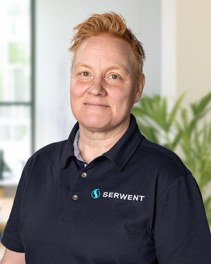 Ann-Christine Ekman, med kort, ljusrött hår, står i ett ljust kontor iklädd en marinblå Serwent-polo. Frodiga växter och stora fönster utgör den livfulla bakgrunden.