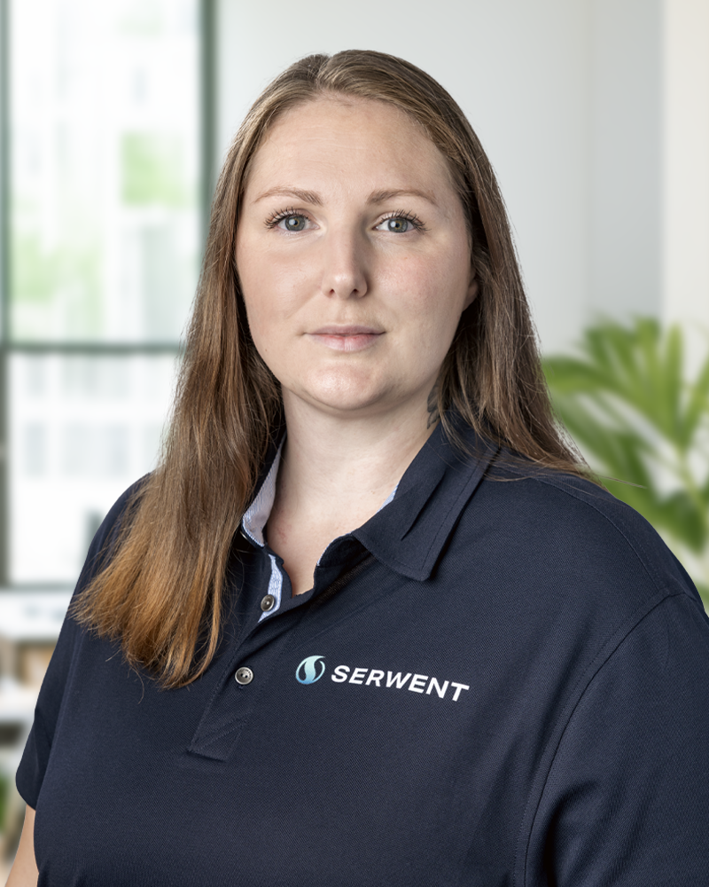 En kvinna med rakt ljusbrunt hår och en marinblå SERWENT-polotröja står i ett ljust kontor med ett stort fönster och en grön växt i bakgrunden.