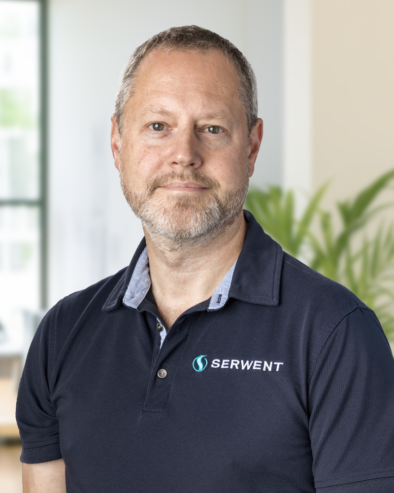 En medelålders man med kort hår och skägg, klädd i en marinblå polotröja med SERWENT-logotypen, står inomhus med suddig grönska och fönster i bakgrunden.