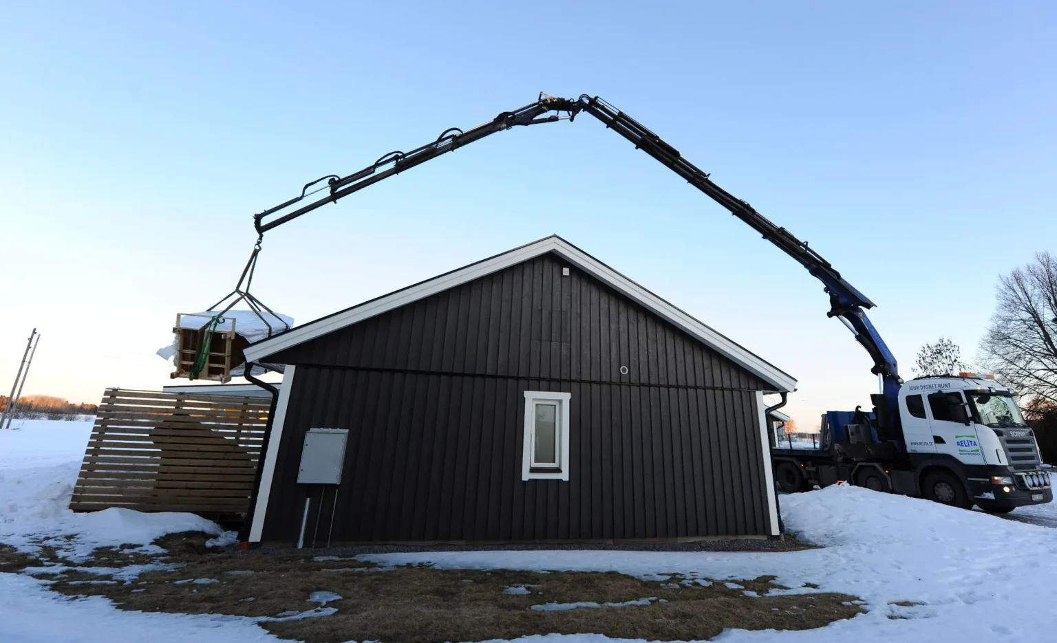 En lastbil med kran levererar byggmaterial över taket på en liten brun byggnad, omgiven av snötäckt mark och klar himmel.