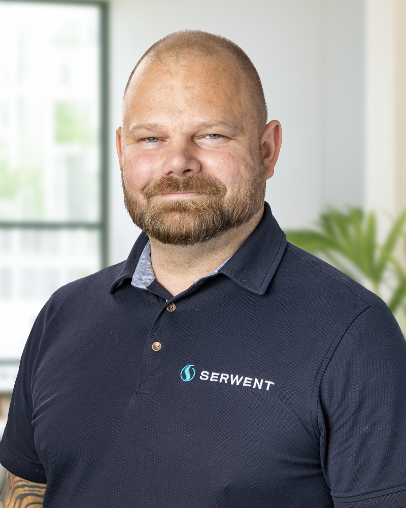 En skäggig man med rakat huvud och en marinblå polotröja med SERWENT-logotypen står inomhus och ler lite. Det finns ett fönster och en grön växt i den suddiga bakgrunden.