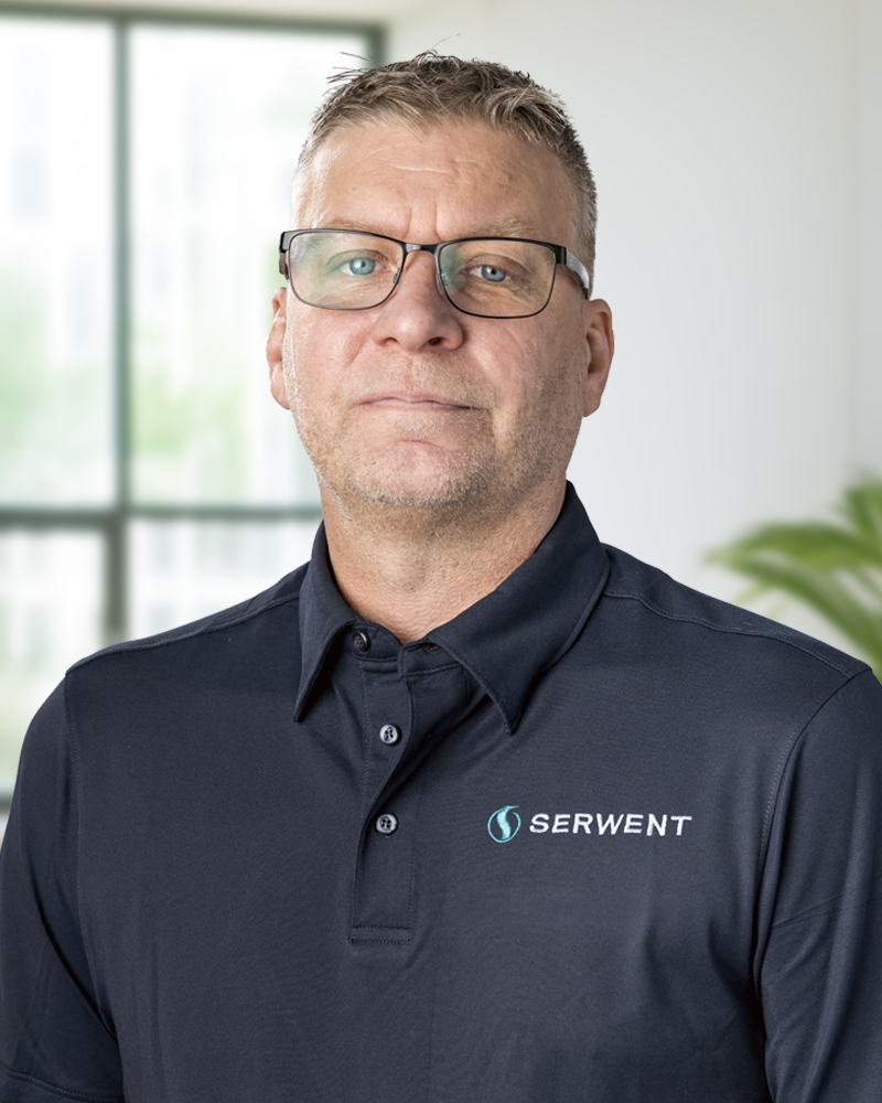 En medelålders man med glasögon och en marinblå polotröja med SERWENT-logotypen står i ett ljust kontor med ett stort fönster och en växt i bakgrunden.