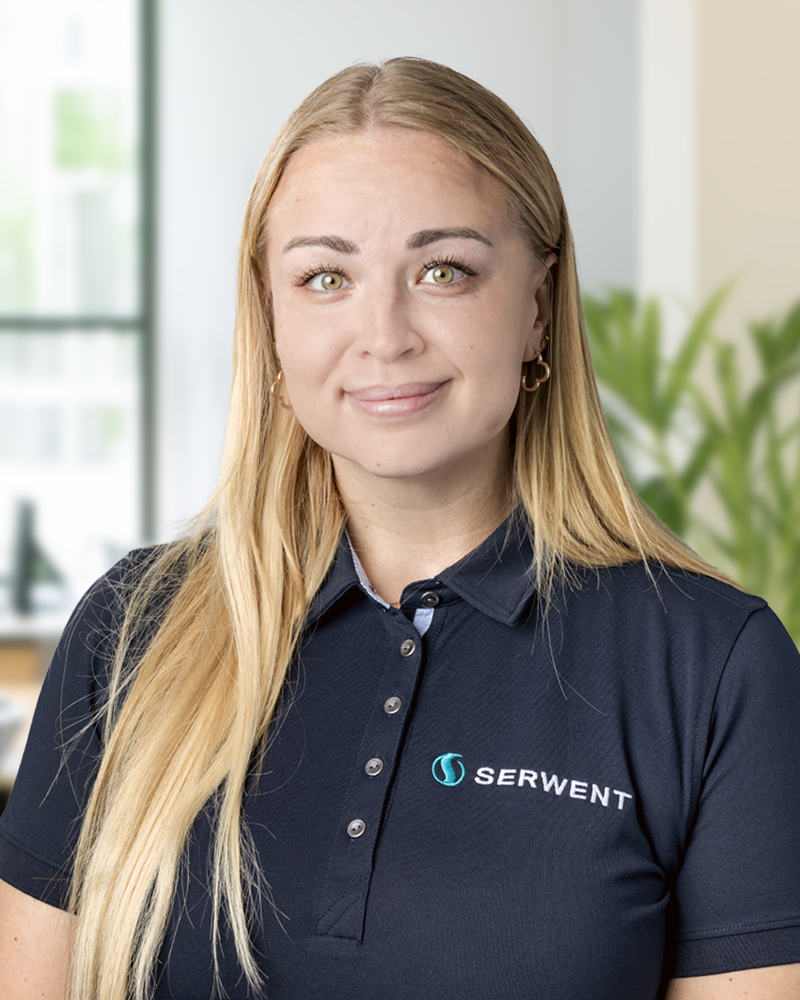 En kvinna med långt blont hår och ljusa ögon, klädd i en marinblå polotröja med SERWENT-logotypen, står inomhus med en suddig kontorsbakgrund och gröna växter.