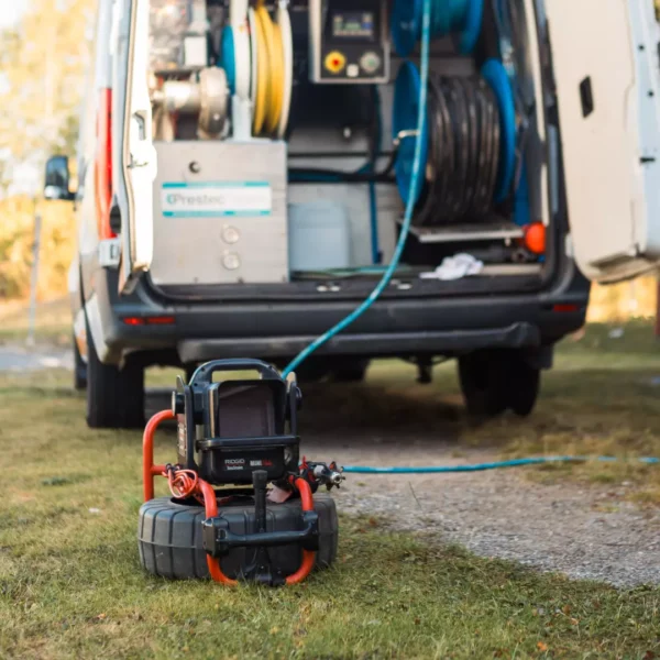 En kamera på hjul med en ansluten kabel placeras på gräsbevuxen mark, vilket leder till den öppna baksidan av en skåpbil fylld med VVS-utrustning, inklusive slangar, inne i en park eller fältområde.
