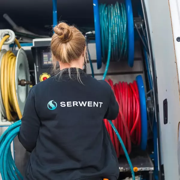 En person med en bullfrisyr, klädd i en svart SERWENT-jacka. Flera spolar av röda, gula och blå kablar syns bakom dem - viktiga verktyg för stamspolning i Stockholm.