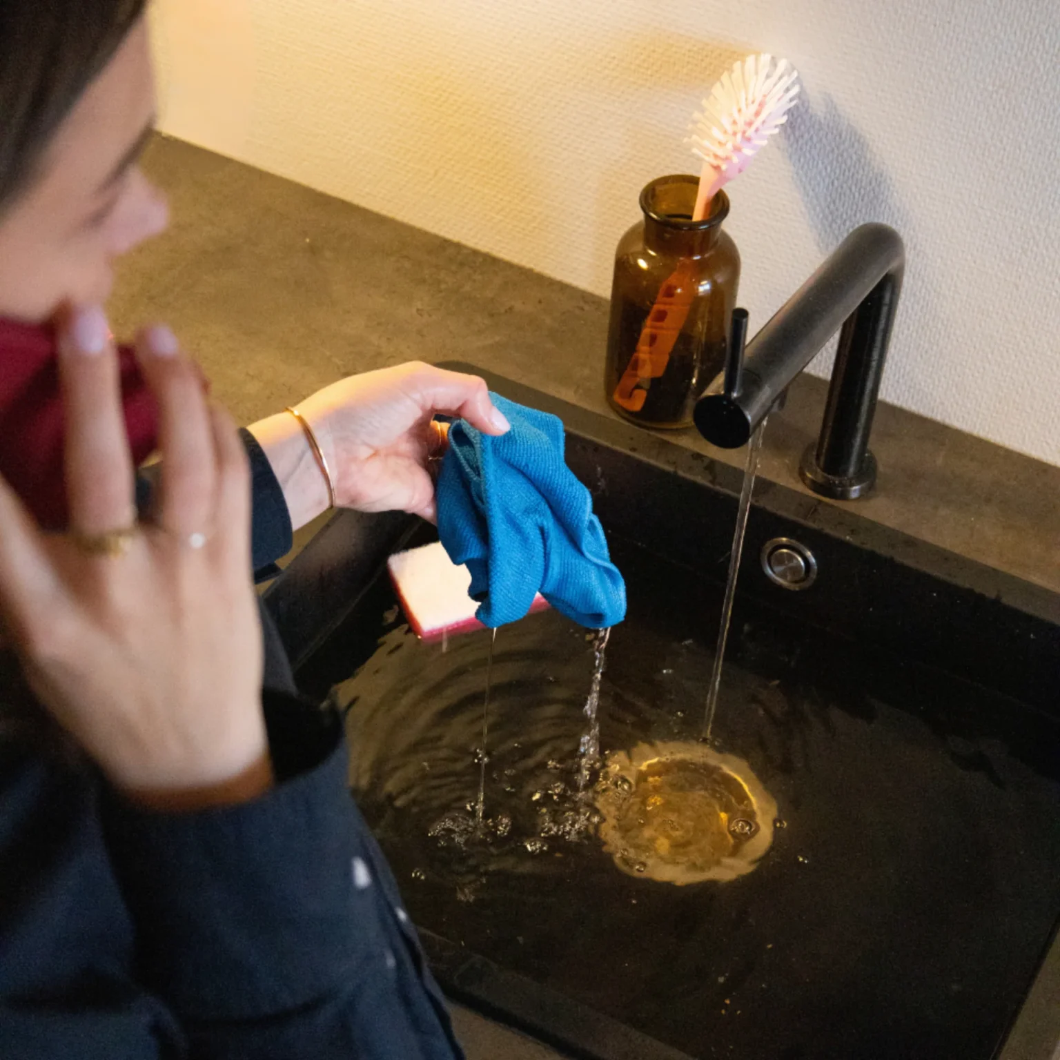 Person som håller en blå trasa under rinnande vatten i ett svart handfat medan han pratar i en smartphone. En borste ligger i en brun vas på diskbänken. Bänkskivan och väggen ger scenen en modern touch.