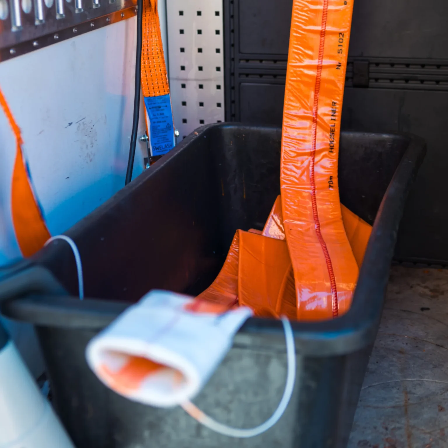 Utrustning för relining ligger organiserad i en svart förvaringslåda, med flera orangea och blåa remmar på en arbetsyta i ett fordon med metallpaneler och perforerad vägg i bakgrunden.