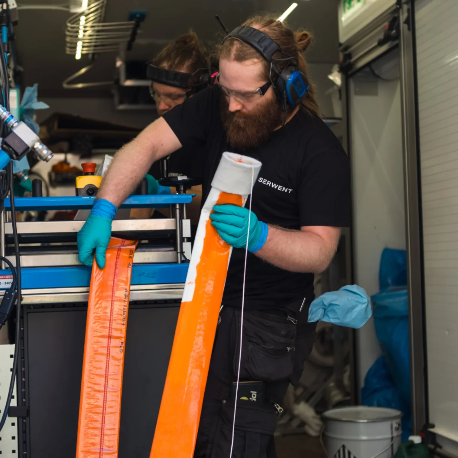 En man som bär skyddsutrustning, inklusive hörselkåpor och handskar, hanterar en orange remsa av material i en verkstad. Han står nära utrustning och hyllor med olika verktyg och förnödenheter i bakgrunden.