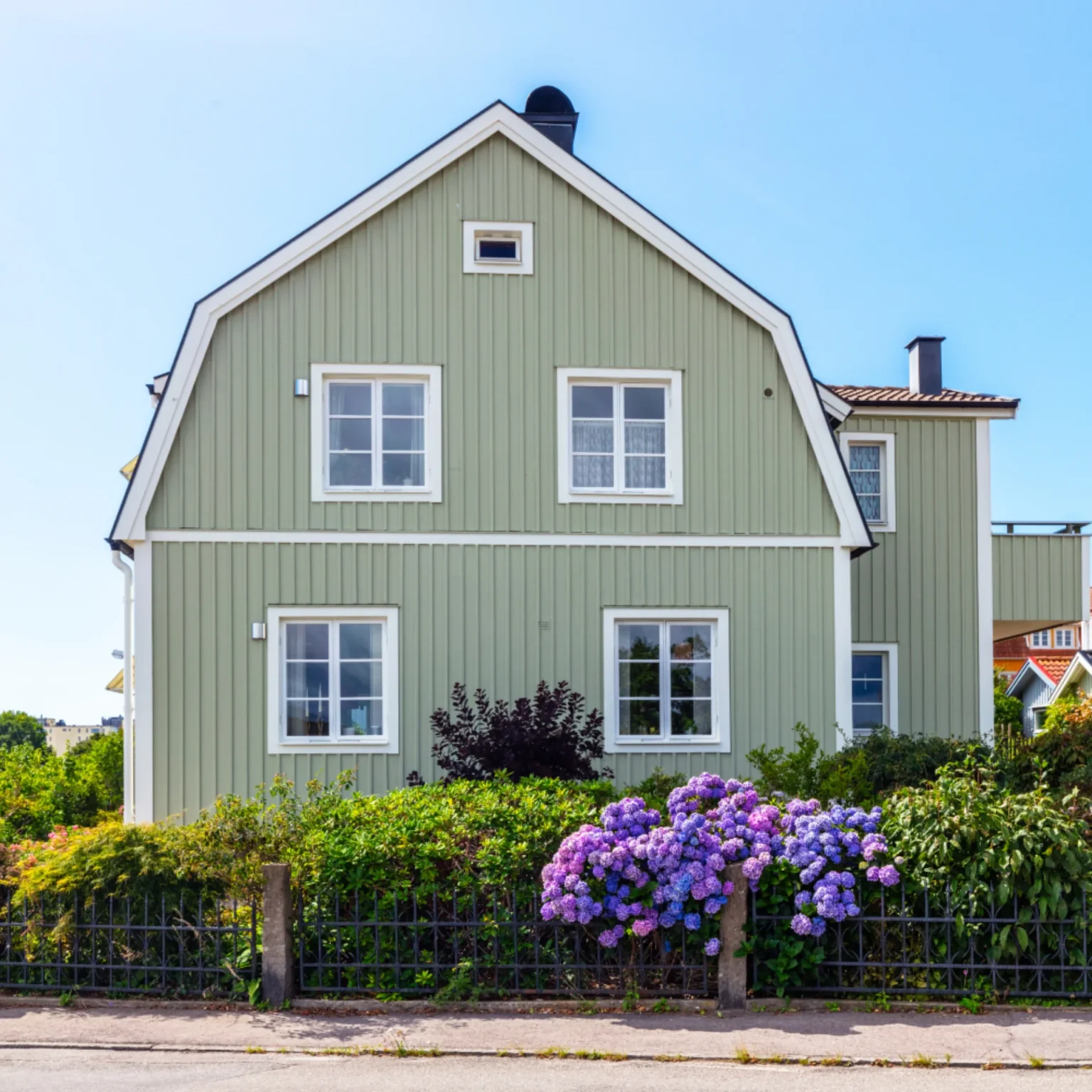 Ett ljusgrönt trähus med vita lister, bakom ett svart staket och färgstarka buskar med lila blommor, har skickligt omklädda fönsterkarmar under en klarblå himmel en solig dag.
