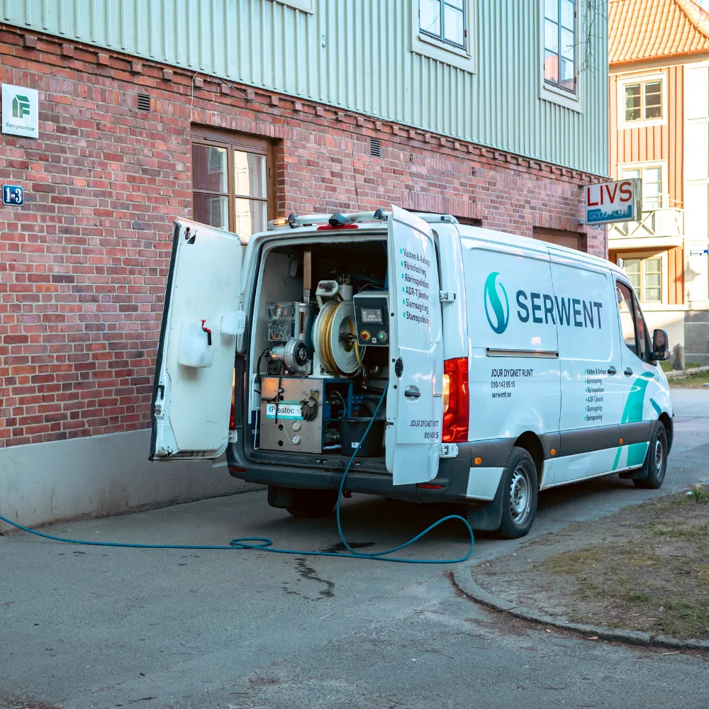 En vit SERWENT servicebil står parkerad bredvid en tegelbyggnad med bakdörrarna öppna, vilket avslöjar rengöringsutrustning och en blå slang som sträcker sig från bilen till marken.