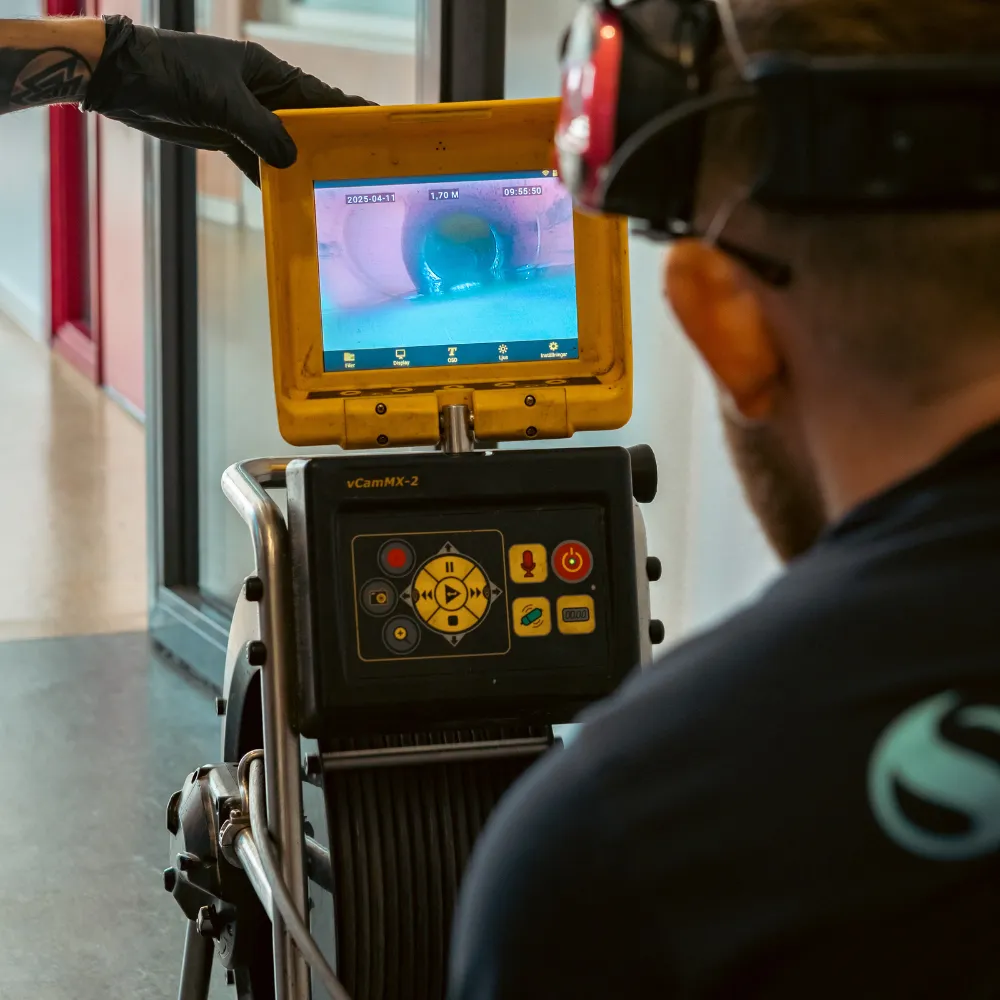 En person som bär handskar och använder en kamerainspektionsenhet tittar på ett rörs insida på en bildskärm och kontrollerar sannolikt blockeringar eller problem i rörsystemet.