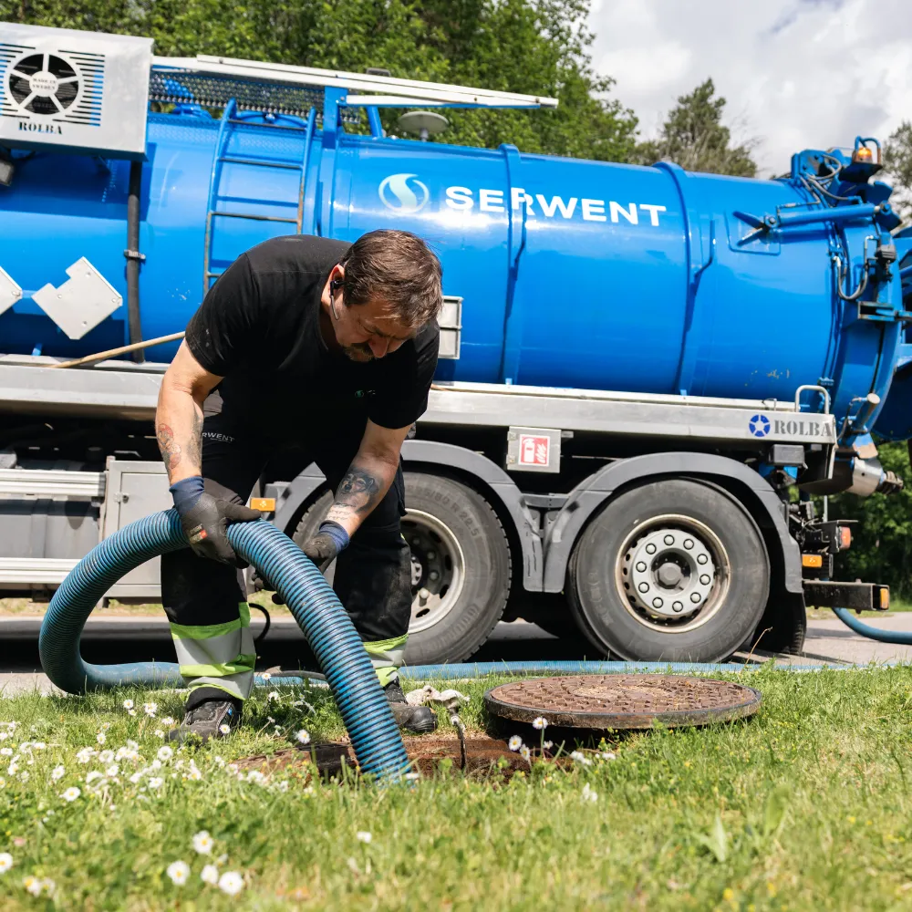 En arbetare i svarta kläder och handskar använder en stor blå slang för att rengöra eller pumpa ur ett manhål, medan en blå septiktankbil märkt SERWENT står parkerad i närheten. Gräs och blommor omger arbetsområdet.