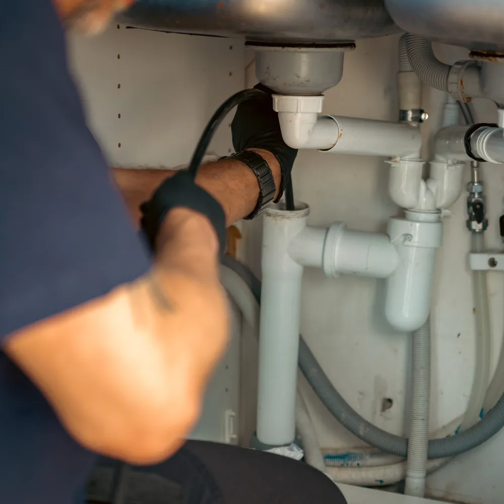En person med handskar använder ett verktyg för att arbeta med vita PVC-rör under en diskbänk och åtgärdar problemet med dålig lukt.