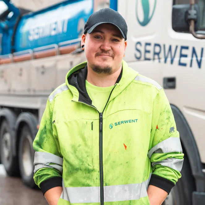En person i neongrön reflekterande arbetsjacka och svart keps står framför en lastbil med texten SERWENT och ler lite. Miljön ser ut att vara utomhus, möjligen på en arbetsplats.