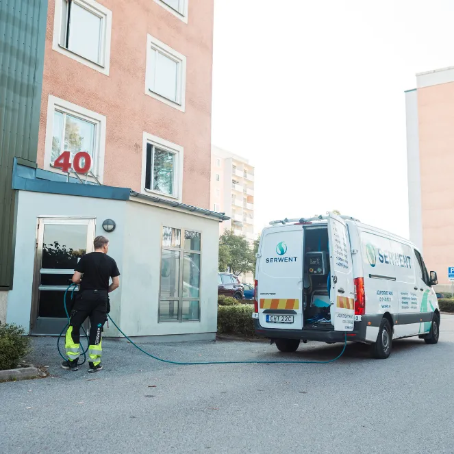 En arbetare i skyddskläder håller i en slang som är ansluten till en vit servicebil märkt Serwent, parkerad utanför ett rosa hyreshus med siffran 40 ovanför entrén.