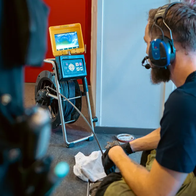 En man med hörlurar och handskar sitter bredvid en monitor som är ansluten till en kabelrulle och inspekterar ett videoflöde från en kamera, troligen för inspektion av rör eller avlopp, med en röd vägg i bakgrunden.