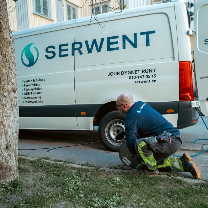 En arbetare i blå skjorta och skyddsbyxor står på knä nära en vit skåpbil märkt Serwent och hanterar en slang eller kabel. Skåpbilen står parkerad på en gata intill ett träd och på sidan står företagets tjänster listade.