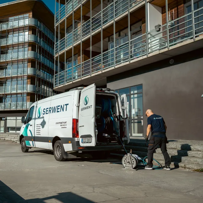 En arbetare står bredvid en vit skåpbil märkt SERWENT utanför ett modernt hyreshus och rullar ut en slang som är ansluten till utrustning i skåpbilen en solig dag.