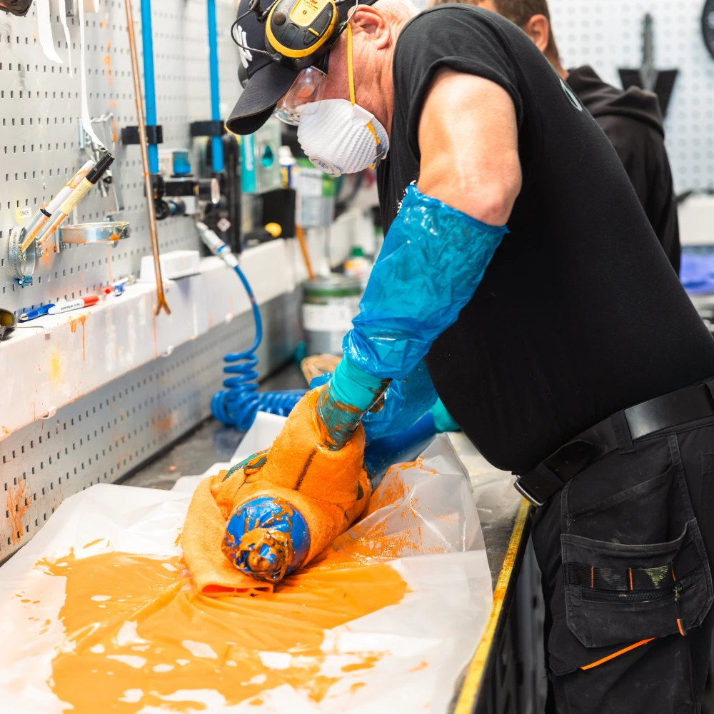 En arbetare som bär skyddsglasögon, handskar, mask och hörselskydd sveper in en industridel med en trasa, medan orange vätska täcker bordet och utrustningen i en verkstadsmiljö.