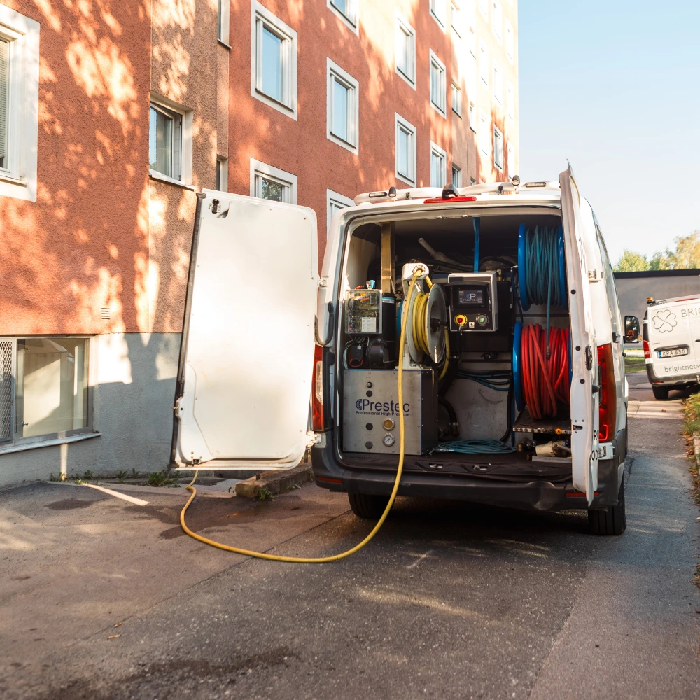 En vit servicebil med öppna bakdörrar parkerad utanför en byggnad. Skåpbilen innehåller slangar, tankar och utrustning för professionell städning eller stambyte-tjänster som "hur ofta görs stambyte?" Solljuset skiner på byggnaden.