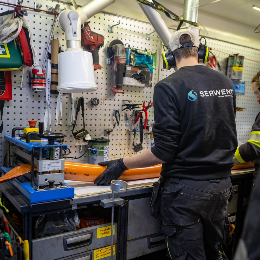 Relining_stockholm_proffs En person iförd skyddsutrustning och en SERWENT-tröja arbetar vid en verktygsfylld arbetsbänk med att hantera en orange slang i en hektisk verkstad med diverse utrustning och verktyg hängande på en pinnbräda.