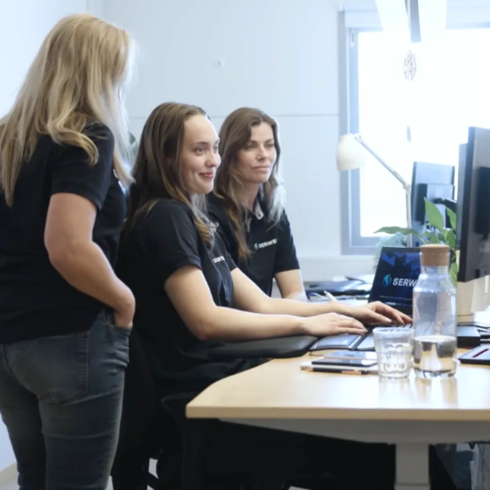 Tre kvinnor i svarta skjortor arbetar tillsammans vid ett skrivbord med datorer i ett ljust kontor. En kvinna sitter och skriver, en annan sitter bredvid henne och den tredje står och tittar på skärmen.
