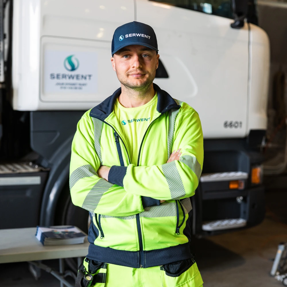 En man i neongul varseluniform och keps står med armarna i kors framför en vit lastbil inne i ett garage i Rotebro. På uniformen och lastbilen syns Serwents logotyp.