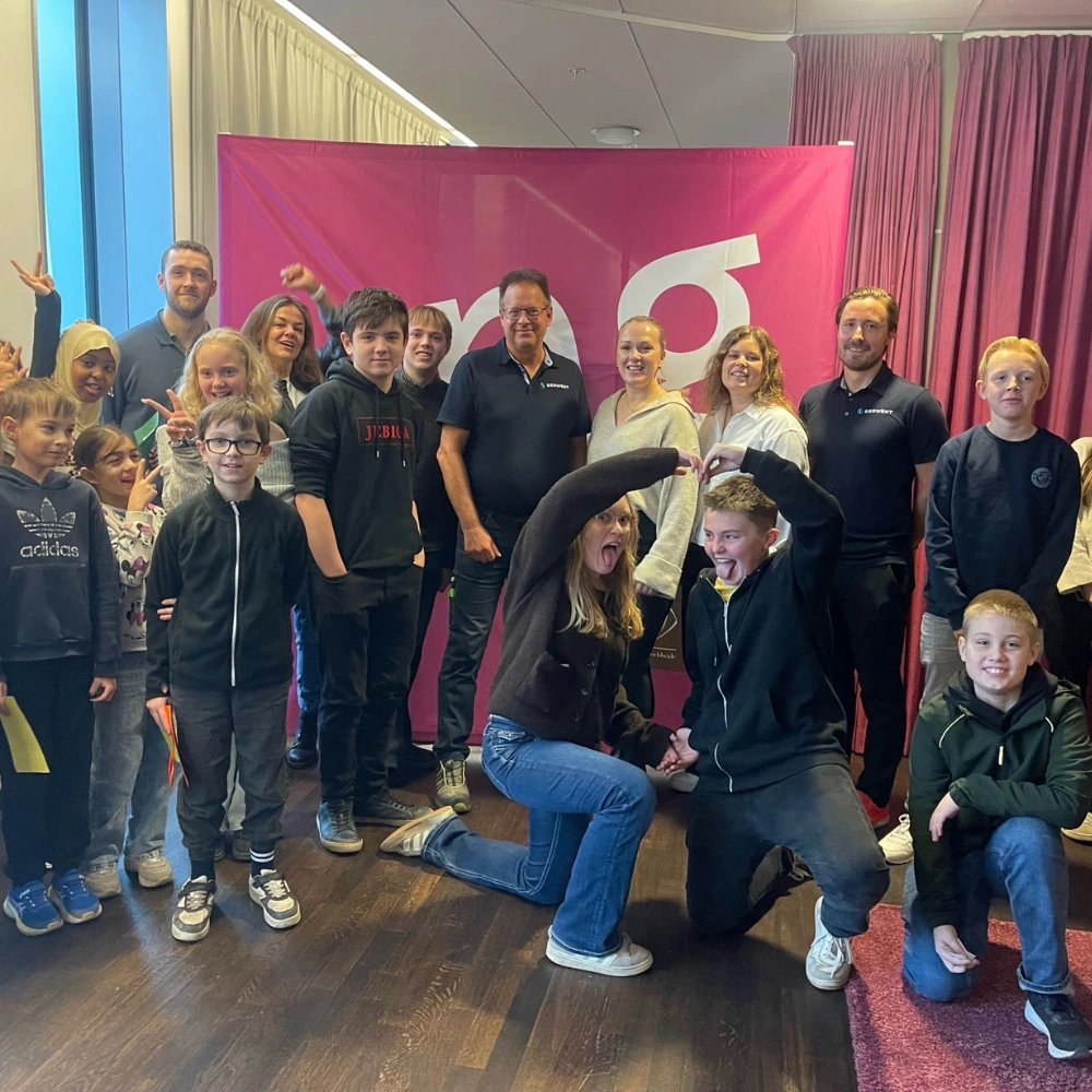 En grupp barn och vuxna poserar och ler tillsammans inomhus framför en stor rosa skylt på Grundskoleutmaningen Serwent. Några visar fredstecken och lekfulla gester, medan andra står eller knäböjer och skapar en glad och livlig stämning.