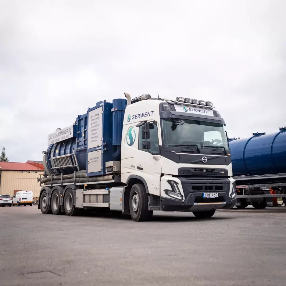 En vit och blå sugbil med märkningen SERVENT står parkerad på en asfalterad tomt nära industribyggnader och en annan blå tankbil under en molnig himmel.