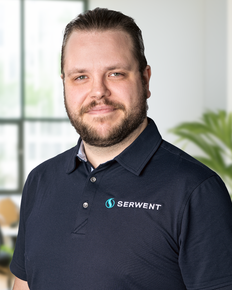En man med skägg och bakåtslickat hår, iklädd en marinblå pikétröja med SERWENT-logotypen, står i ett modernt, ljust kontor med stora fönster och en grön växt i bakgrunden.