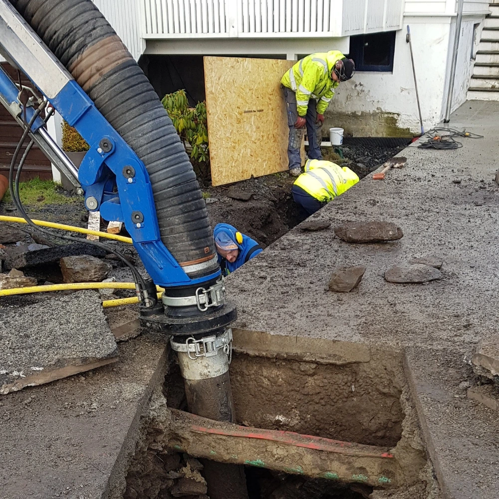 Två byggnadsarbetare i skyddsutrustning reparerar rör i ett uppgrävt område nära ett hus. Tunga maskiner med en stor slang används för att gräva upp jord. Trasiga trottoarplattor ligger runt om på arbetsplatsen.
