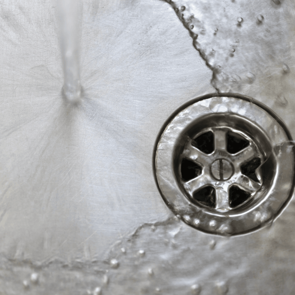 Vatten rinner ner i en diskho i rostfritt stål nära ett metallavlopp, vilket skapar ett cirkulärt stänkmönster och små vattendroppar - tecken på att stopp i avloppet snart kan inträffa om flödet blockeras.