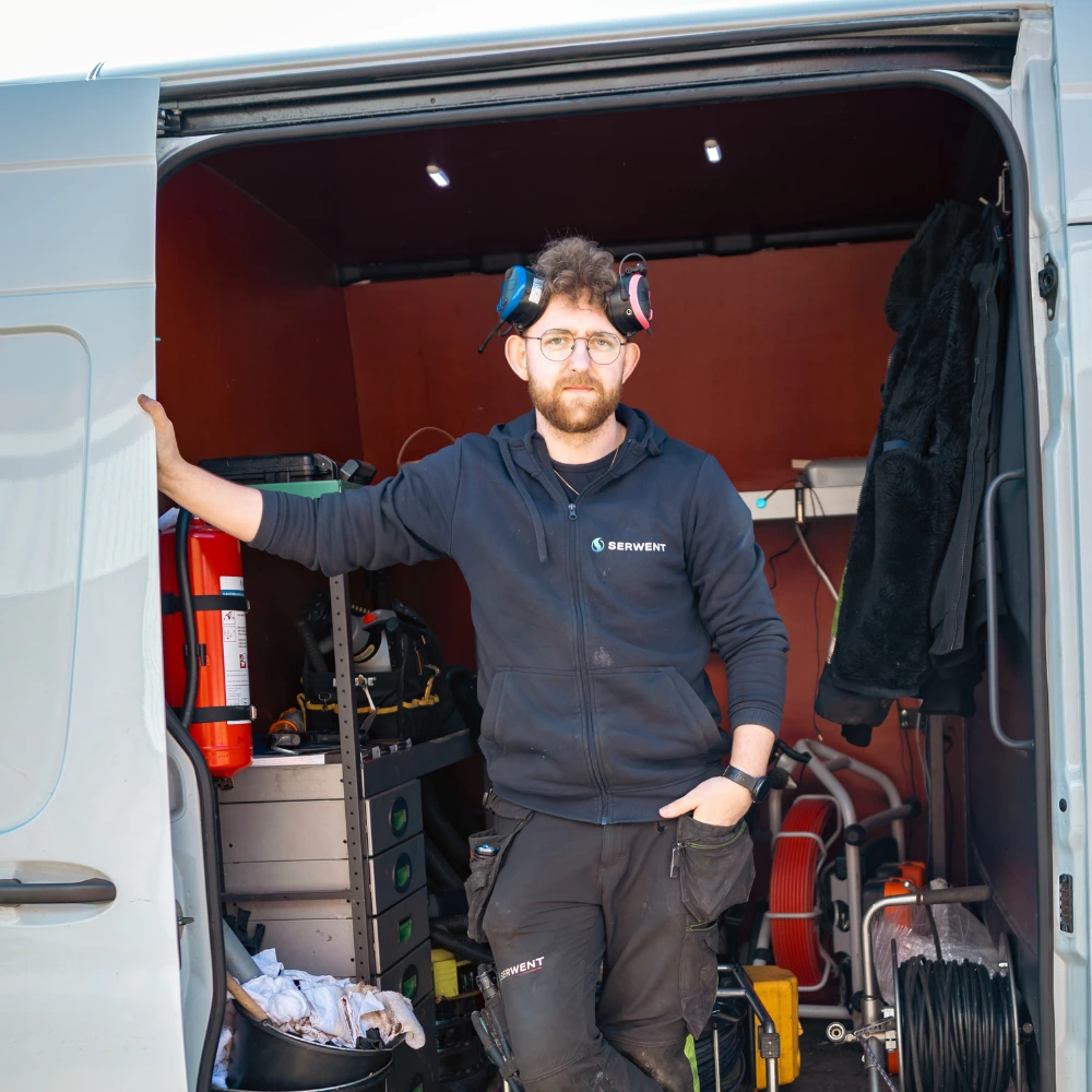 En man med glasögon och hörlurar runt halsen står i den öppna sidodörren på en arbetsbil, omgiven av verktyg och utrustning, iklädd en svart huvtröja och arbetsbyxor.