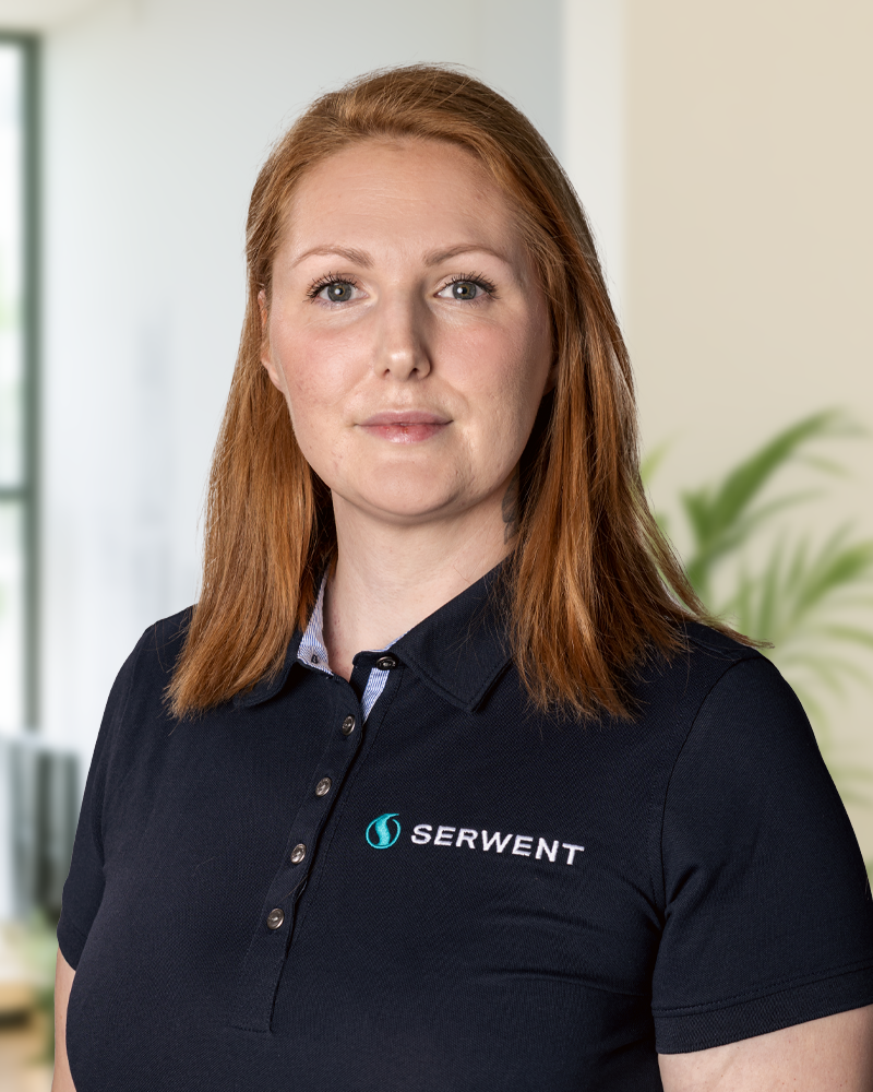 En kvinna med långt rött hår och svart polotröja med SERWENT-logotypen står inomhus och tittar in i kameran. I bakgrunden syns en suddig växt och ett fönster - ett kontor där Patrik Gustavssons team utmärker sig.
