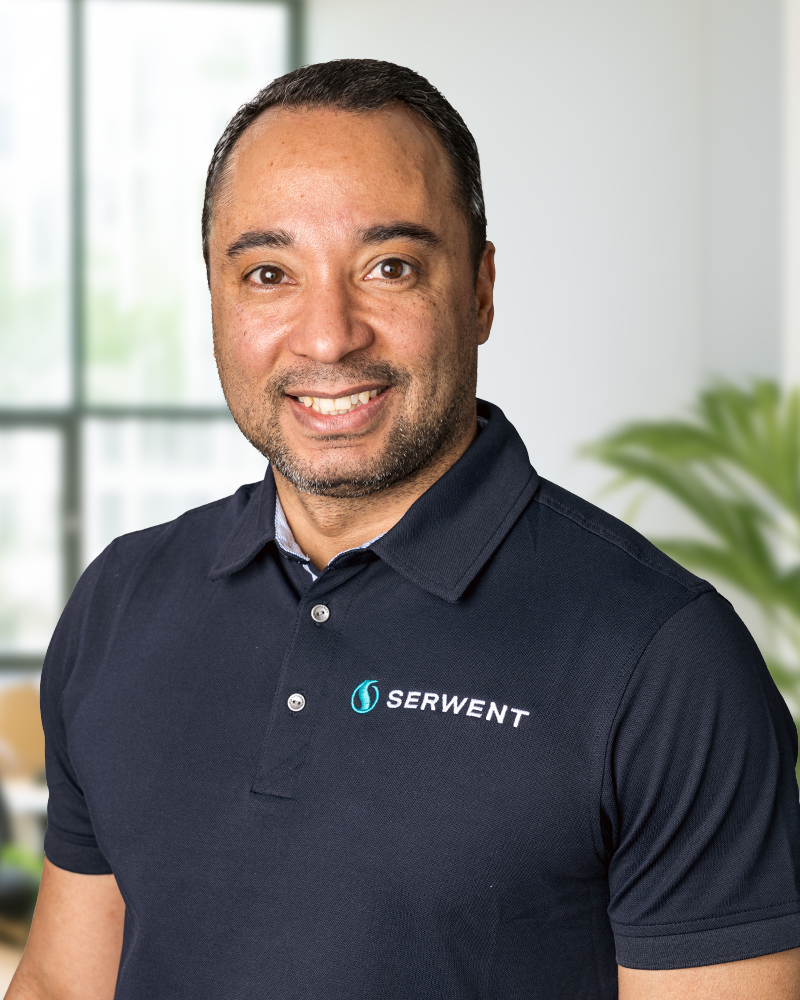 En leende man i marinblå polotröja med SERWENT-logotypen står i ett modernt kontor med stora fönster och en grön växt i bakgrunden. Det här är Patrik Gustavsson.