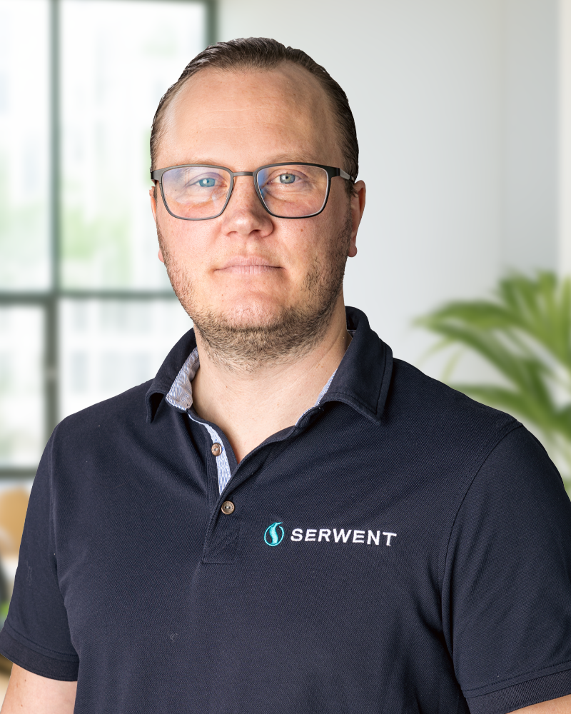 Patrik Gustavsson, en man med kort hår och glasögon, står inomhus iklädd en marinblå SERWENT-polotröja, inramad av stora fönster och gröna växter i den suddiga bakgrunden.