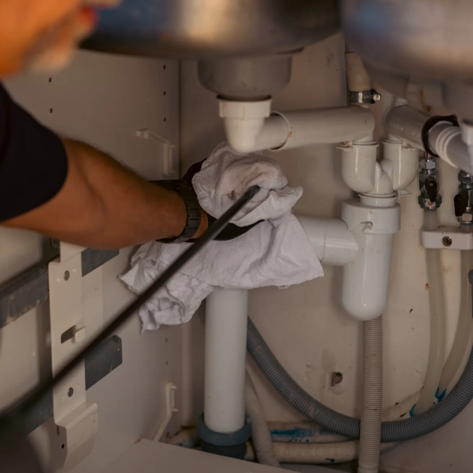 En person använder en rörslang för att rensa rör under ett handfat och håller en vit trasa runt avloppsröret som skydd. Under diskbänken syns olika plaströr, slangar och anslutningar.