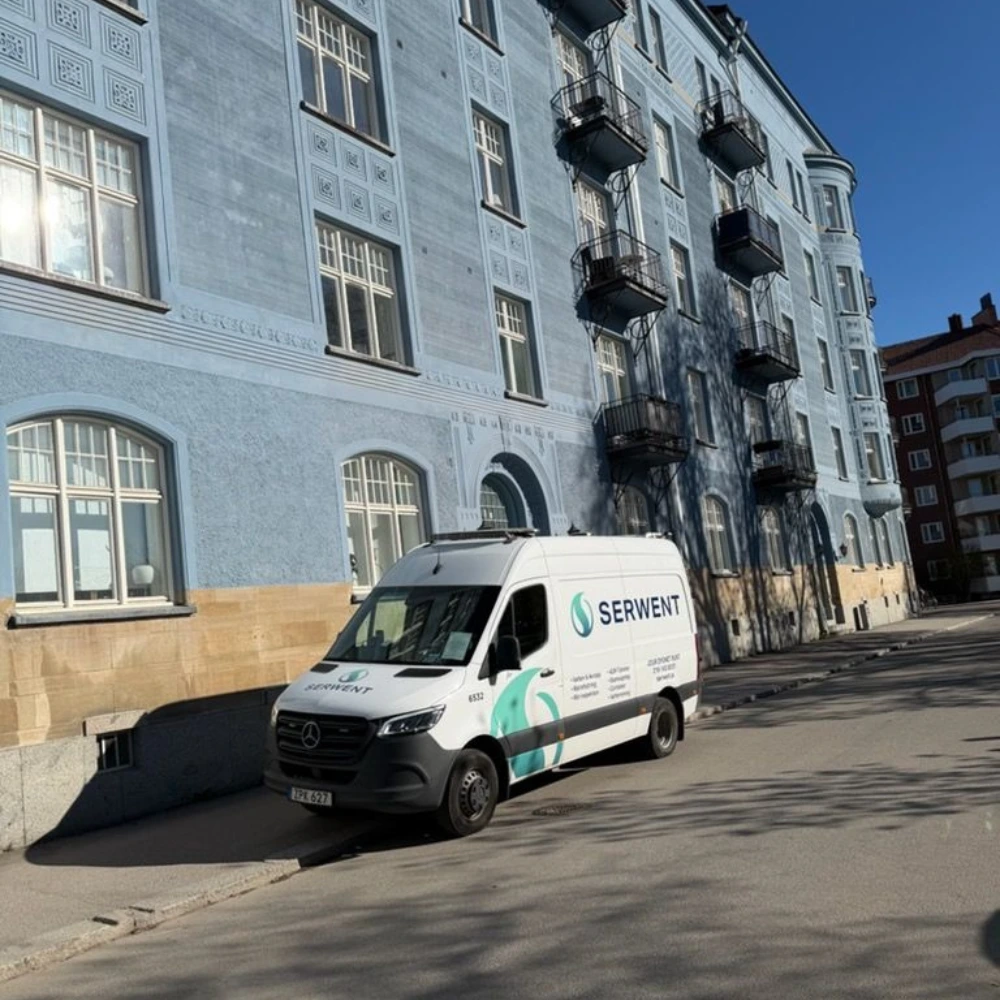 En vit skåpbil med logotypen SERVENT, specialiserad på stamspolning i Uppsala, står parkerad på en solig gata bredvid en stor blå byggnad med balkonger och välvda fönster. Skuggor från närliggande träd faller över trottoaren och byggnaden.