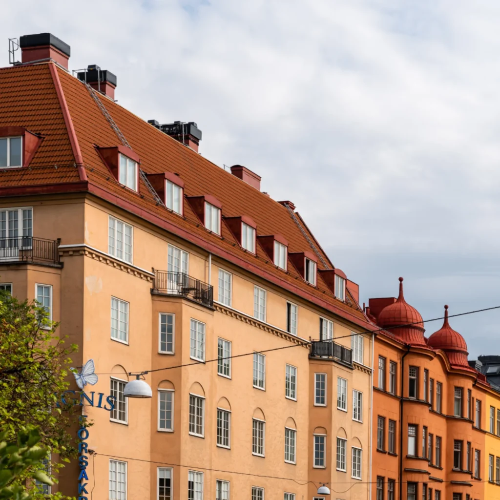 En rad färgglada flerfamiljshus med röda tegeltak, orange och beige fasader och rundade torn under en delvis molnig himmel. Träd och gatlyktor syns i förgrunden.