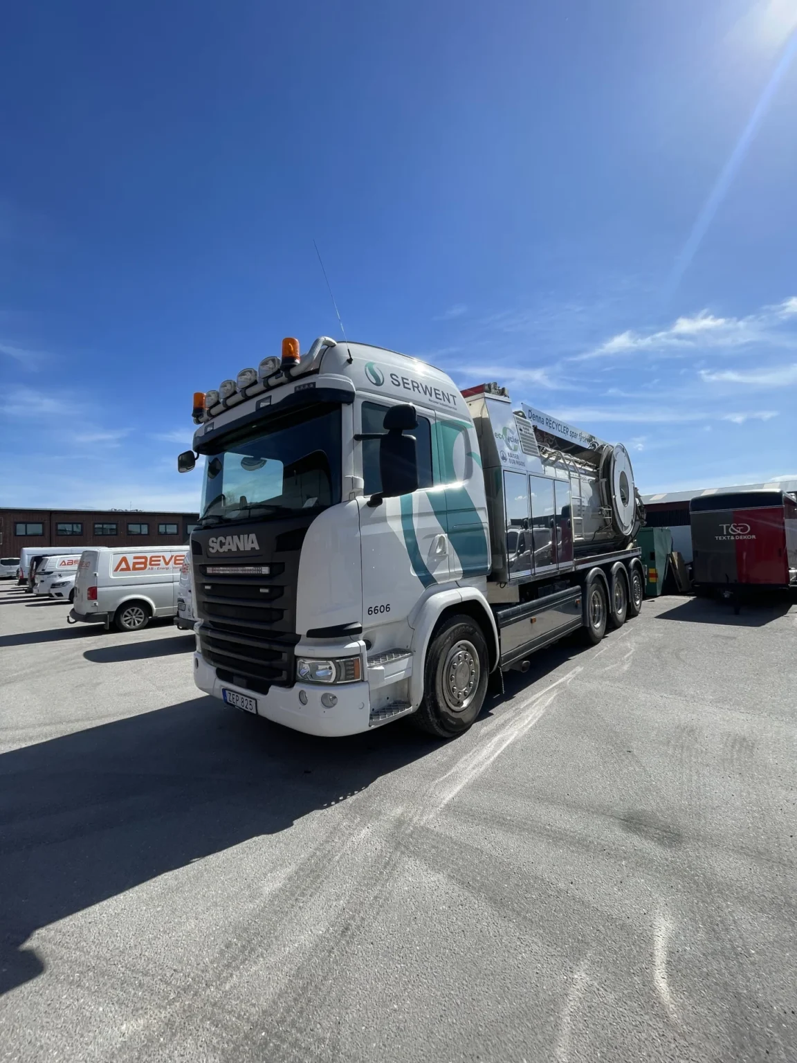 En stor vit Scania lastbil med Vattenreningssystem står parkerad på en parkeringsplats under en solig blå himmel, med ett släp och andra fordon synliga i bakgrunden.