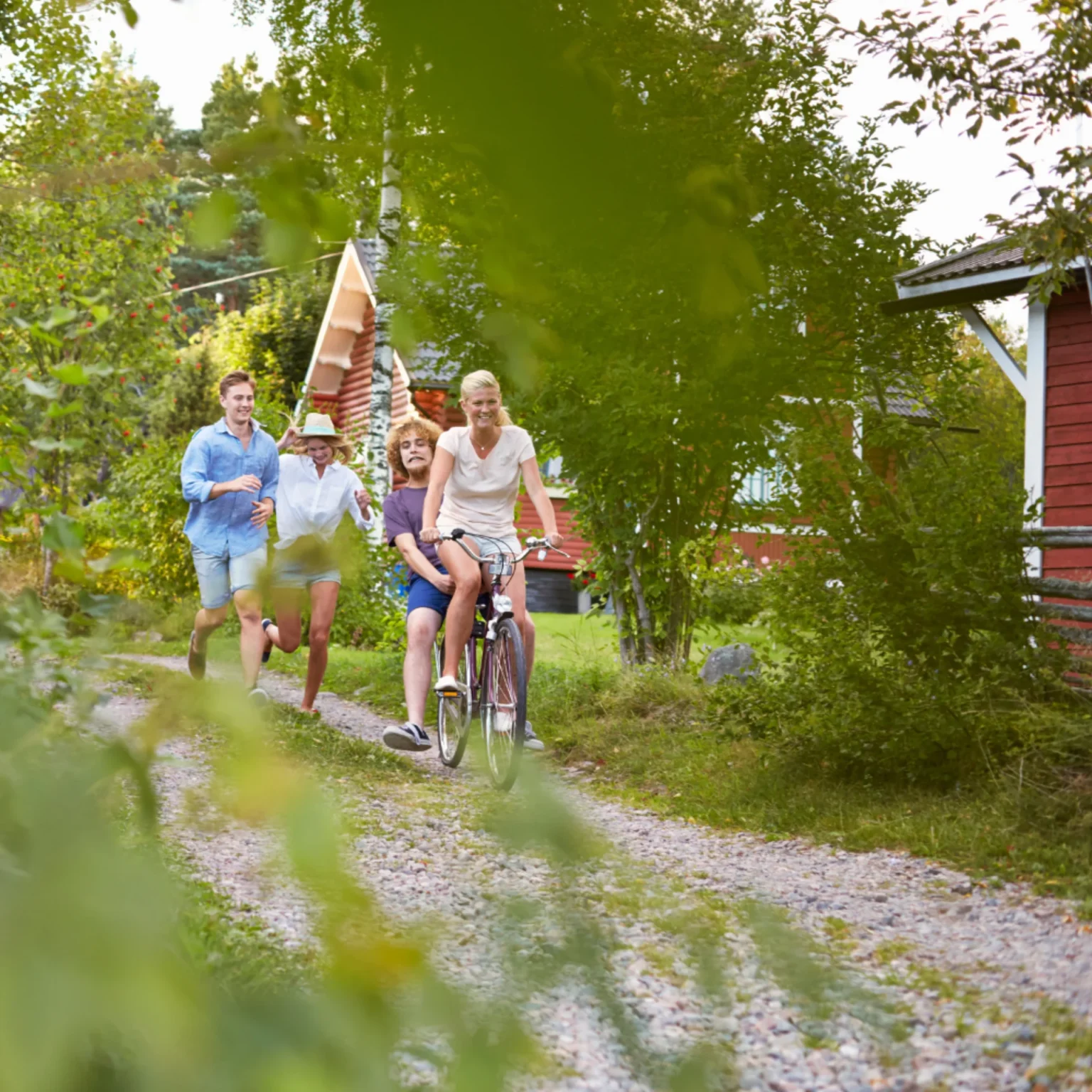 Fyra personer njuter av en sommardag utomhus; en cyklar med ett barn på styret medan två andra springer längs en grusväg nära röda trästugor och pratar om senaste avloppsproblemet, omgivna av gröna träd och gräs.