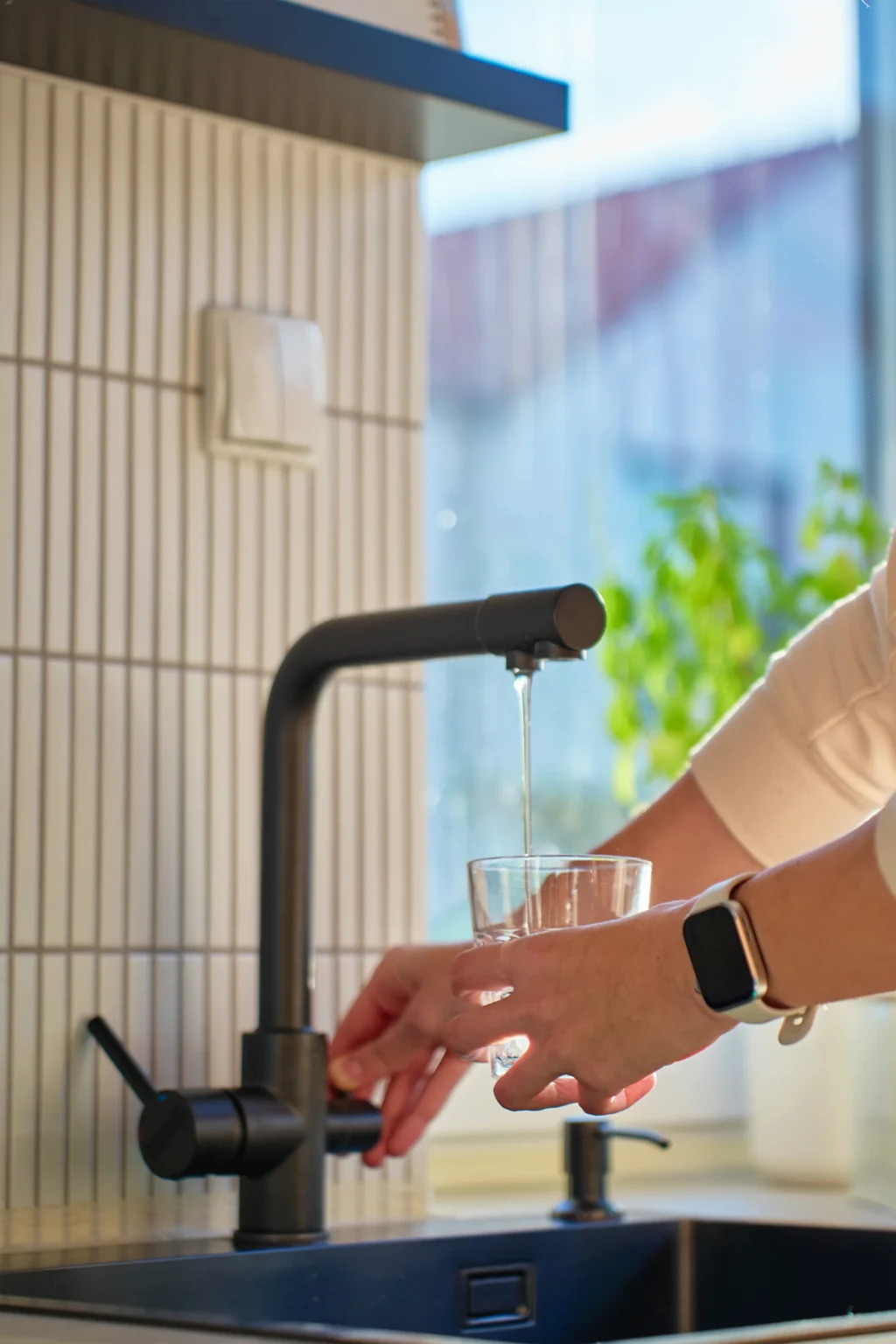 En person med en smartklocka fyller ett glas med vatten från en modern svart köksblandare med avancerad vattenrening, med solljus som strömmar in genom ett fönster i bakgrunden.