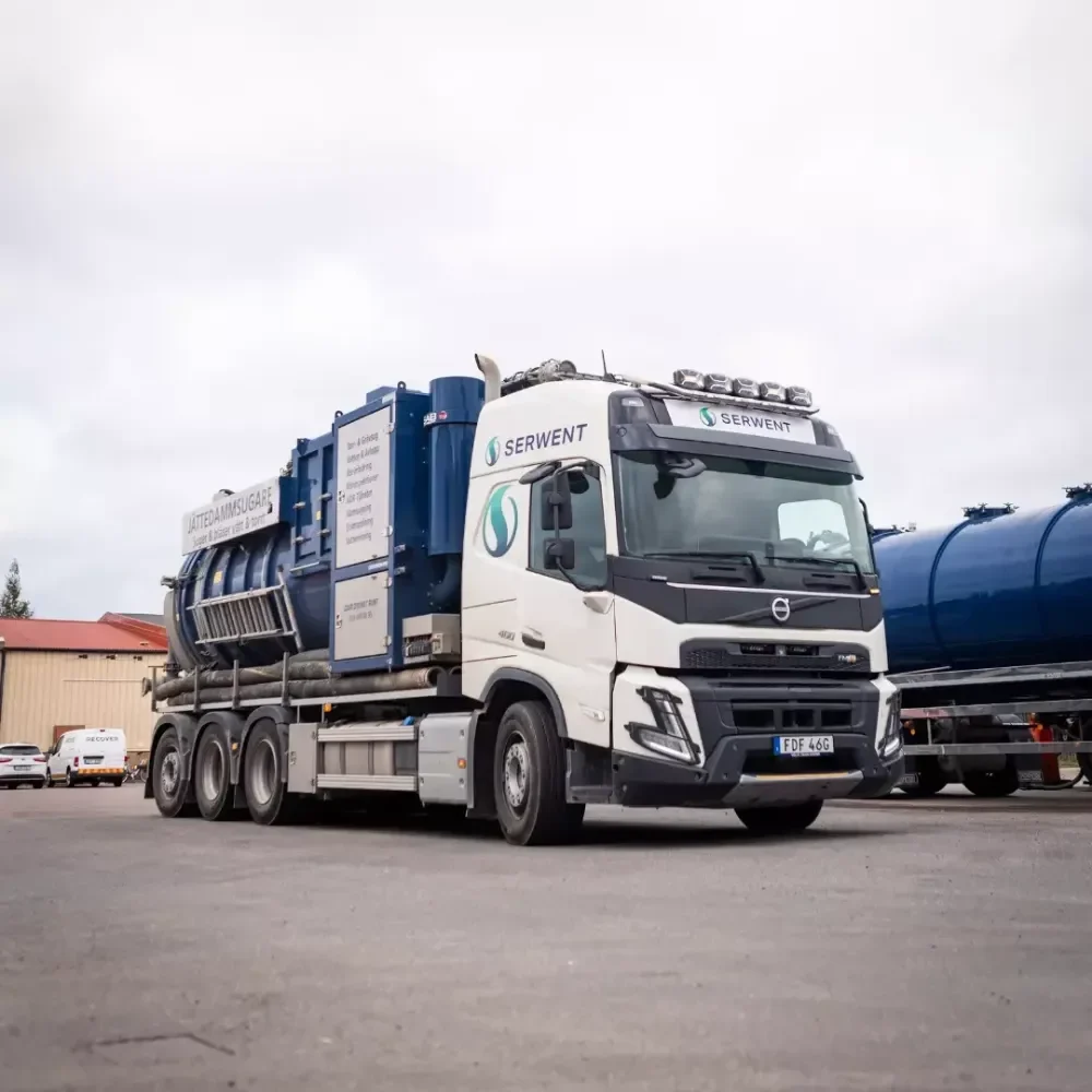 Serwent Stockholm En vit och blå SERVENT sugbil, med ADR-tjänster Stockholm, står parkerad på en asfalterad tomt med industribyggnader och en annan blå tankbil i bakgrunden under en molnig himmel.