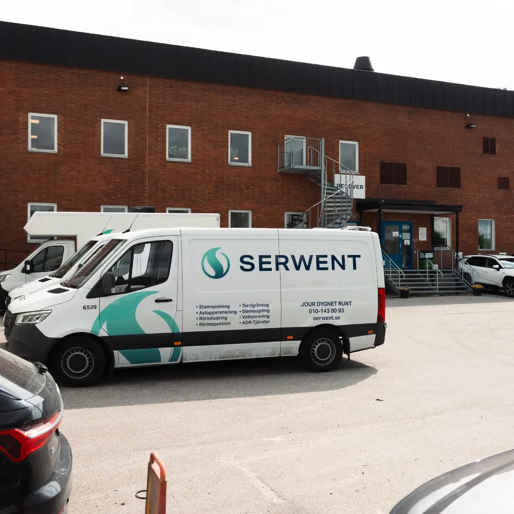 Serwent Stockholm kontor En vit skåpbil märkt SERWENT, känd för ADR-tjänster Stockholm, står parkerad utanför en industribyggnad i tegel med metalltrappor som leder till en övre dörr. Flera andra fordon är också parkerade i närheten på tomten.