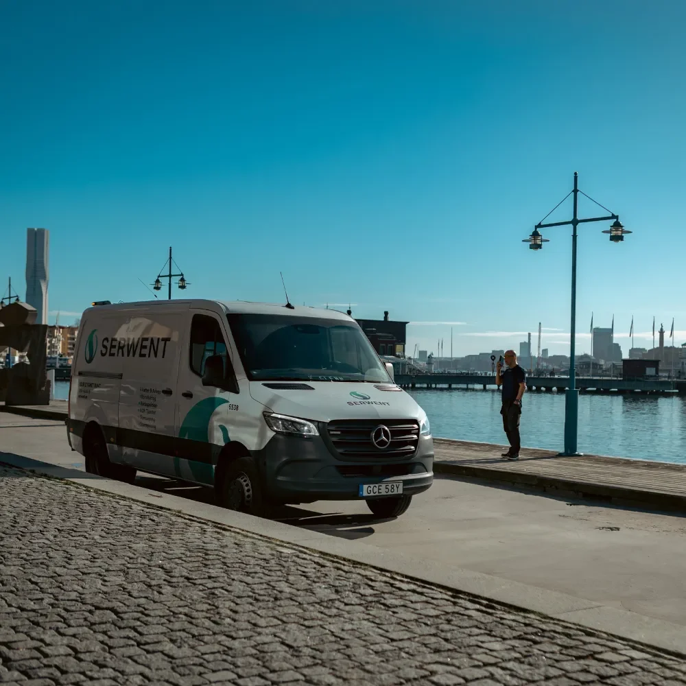 Serwent Göteborg En vit skåpbil från Serwent Göteborg står parkerad vid vattnet en solig dag. En person står i närheten, med byggnader och lyktstolpar som kantar vattnet under den klarblå himlen.