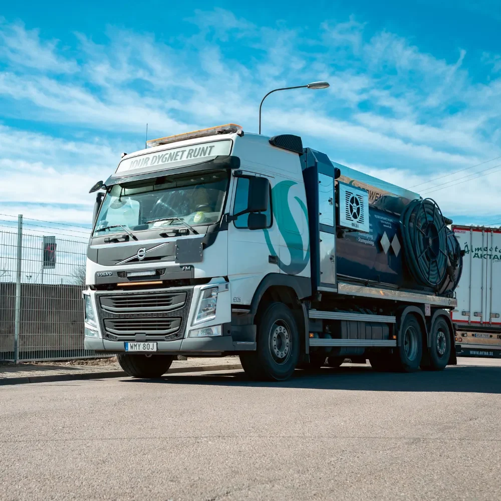 Fordorn - Serwent En stor vit och blå Volvo lastbil med Serwent Uppsala utrustning på flaket är parkerad på en solig gata nära ett staket och en annan lastbil, under en ljusblå himmel med spridda moln.