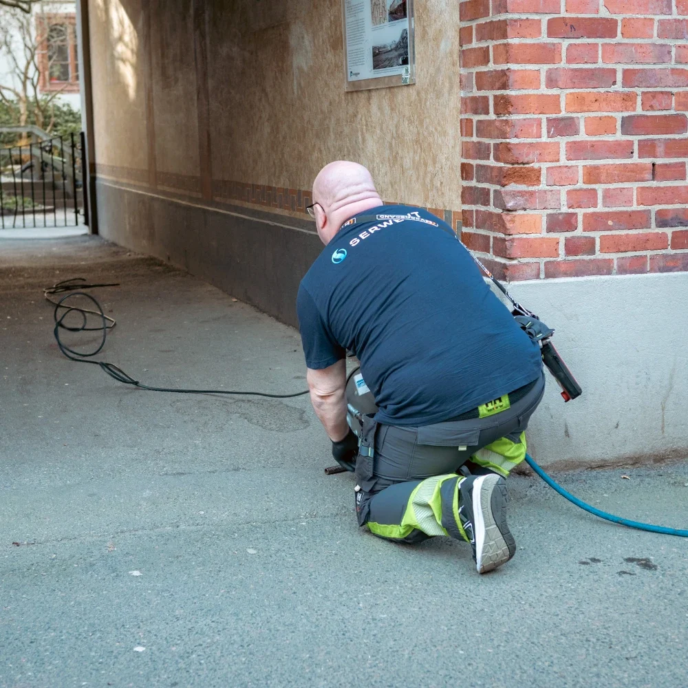 lägenhet stamspolning En person i arbetskläder och skyddsutrustning står på knä på marken i en gränd och använder elverktyg med kablar och slangar, vänd bort från kameran nära en tegel- och stuckaturvägg.