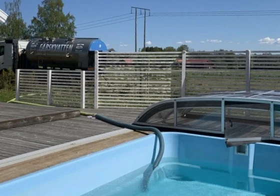 En pool som fylls med vatten med hjälp av en stor slang ansluten till en tankbil. Bakgrunden inkluderar ett trädäck, ett staket, träd och en klarblå himmel.