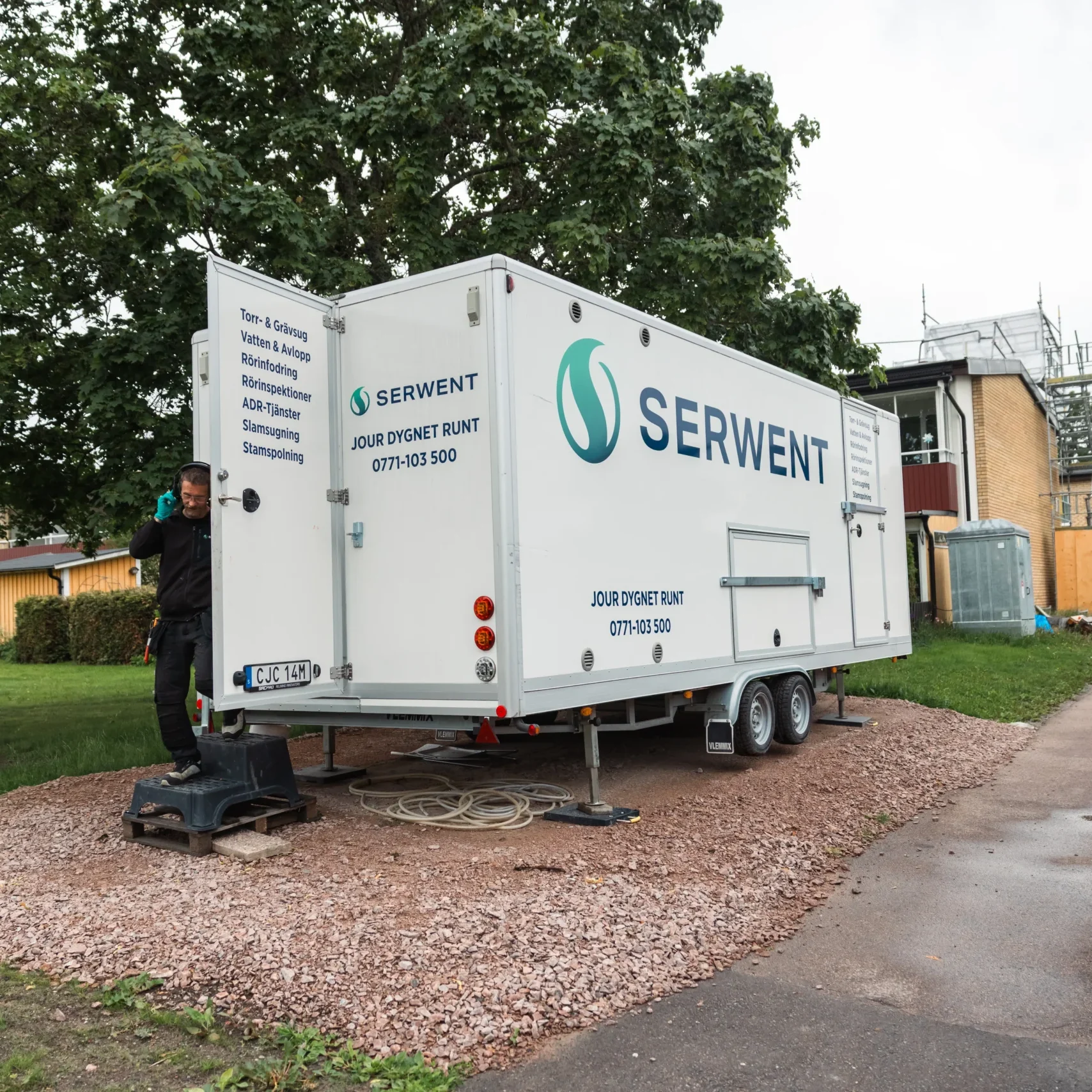 Serwent Mälardalen En person står bredvid en stor vit släpvagn märkt SERWENT, parkerad på en grusplan nära en gräsmatta med träd och bostadshus i bakgrunden.